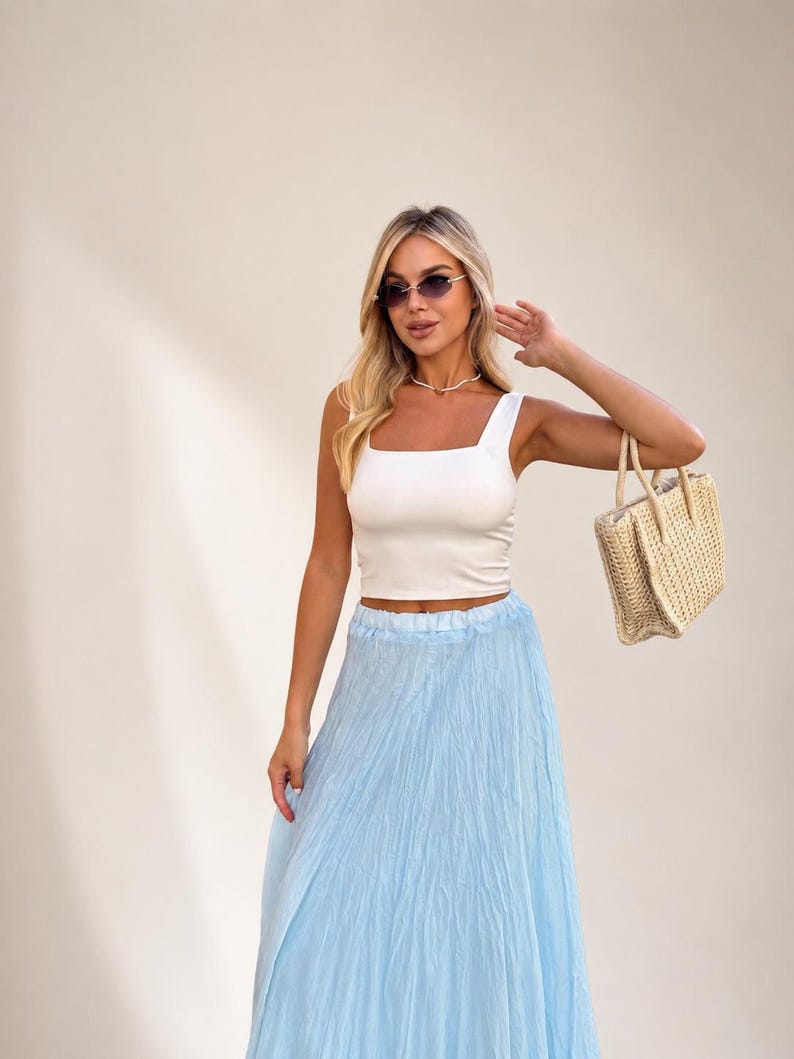 May include: A white sleeveless crop top with a square neckline paired with a light blue crinkled maxi skirt. The outfit is accessorized with a woven straw handbag, a delicate necklace, and dark-tinted sunglasses with thin gold-toned frames.