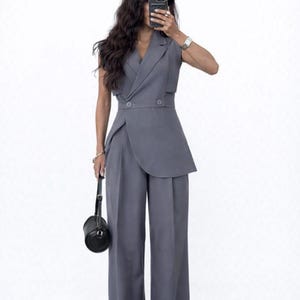May include: A grey sleeveless blazer and wide-leg trousers ensemble. The blazer has a wrap-style design with two buttons. A small black handbag is held in the left hand. The outfit is a solid grey colour.