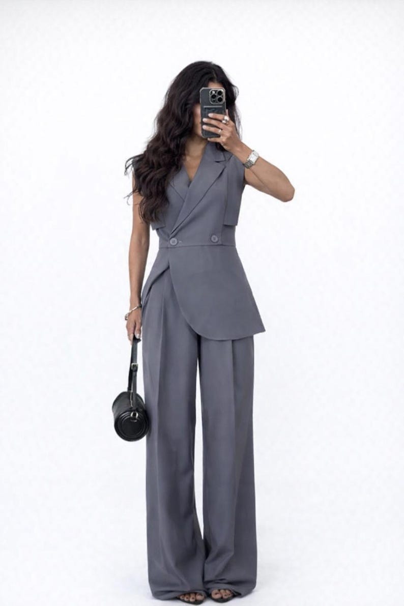 May include: A grey sleeveless blazer and wide-leg trousers ensemble. The blazer has a wrap-style design with two buttons. A small black handbag is held in the left hand. The outfit is a solid grey colour.