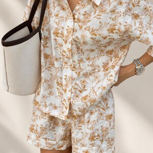 Puede incluir: Un conjunto de camisa y pantal&oacute;n corto con estampado floral blanco con un bolso tote a juego. La camisa es abotonada con mangas cortas. El pantal&oacute;n corto tiene un corte relajado. El bolso tote es de color crema con asas marrones.