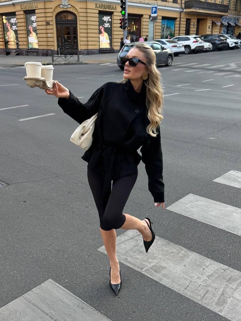 May include: A black belted jacket with long sleeves, black leggings, and black pointed-toe slingback heels. The outfit includes a white handbag, black sunglasses, and a cardboard carrier holding two white coffee cups with lids.