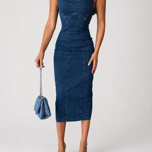 May include: A dark blue denim midi dress featuring a square neckline, thick shoulder straps, and vertical seam detailing along the bodice and skirt. The dress is paired with a small quilted denim handbag with a silver chain and blue denim mules.