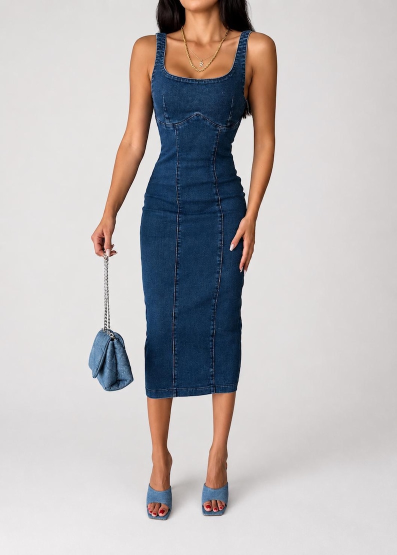 May include: A dark blue denim midi dress featuring a square neckline, thick shoulder straps, and vertical seam detailing along the bodice and skirt. The dress is paired with a small quilted denim handbag with a silver chain and blue denim mules.