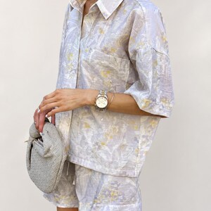 Puede incluir: Un conjunto de dos piezas a juego con una camisa con botones y pantalones cortos. El conjunto es de color lavanda claro con un estampado floral en amarillo y blanco. La camisa tiene un bolsillo en el pecho. Se sostiene un bolso gris en la mano.