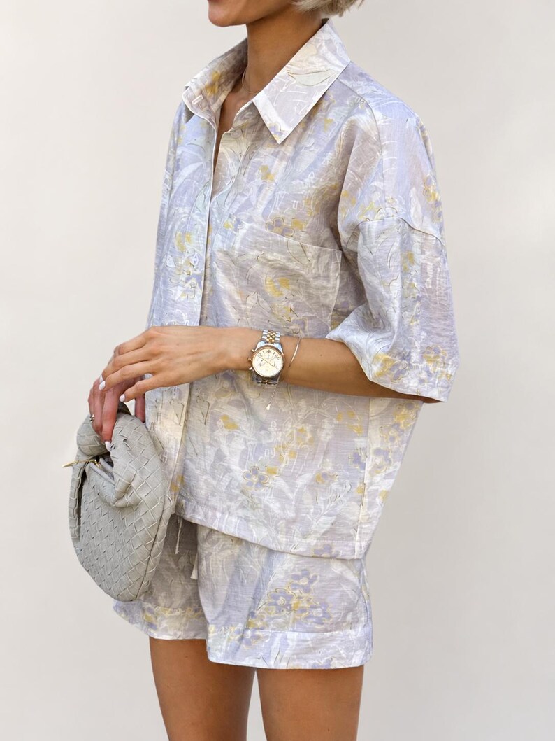 Puede incluir: Un conjunto de dos piezas a juego con una camisa con botones y pantalones cortos. El conjunto es de color lavanda claro con un estampado floral en amarillo y blanco. La camisa tiene un bolsillo en el pecho. Se sostiene un bolso gris en la mano.