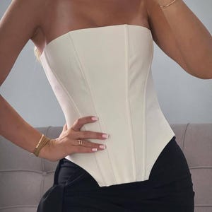 May include: Cream-colored strapless corset top with vertical seams. The top is paired with black high-waisted pants. The corset has a structured, fitted design. The overall look is chic and modern.
