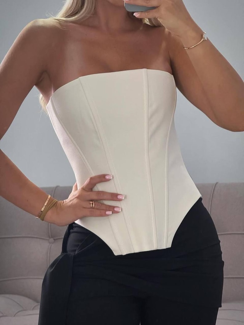 May include: Cream-colored strapless corset top with vertical seams. The top is paired with black high-waisted pants. The corset has a structured, fitted design. The overall look is chic and modern.