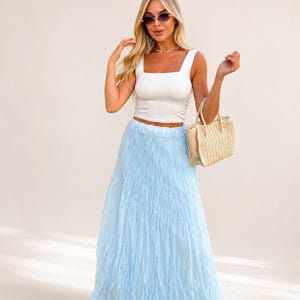 May include: A white sleeveless crop top with a square neckline paired with a long, light blue, crinkled texture maxi skirt. A small, rectangular, beige woven straw handbag with two top handles is held at the side of the skirt.