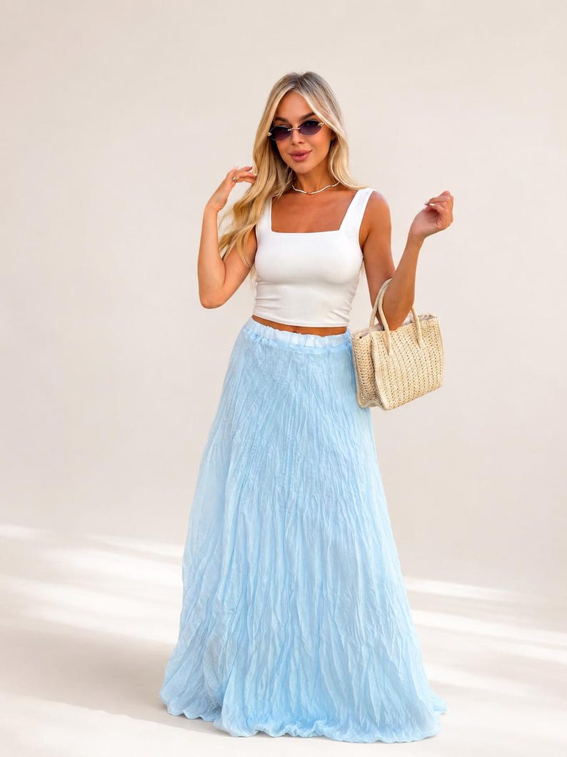 May include: A white sleeveless crop top with a square neckline paired with a long, light blue, crinkled texture maxi skirt. A small, rectangular, beige woven straw handbag with two top handles is held at the side of the skirt.