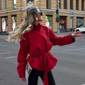 May include: A vibrant red jacket featuring a high collar, long sleeves, and a matching fabric tie belt at the waist. The jacket has a structured fit with a flared hemline and is paired with solid black leggings. The garment is worn with dark sunglasses.
