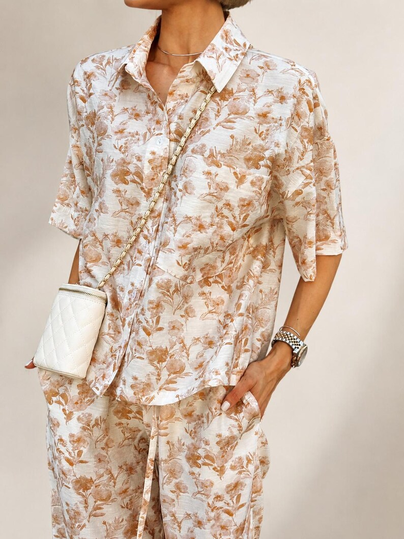 Puede incluir: Conjunto de dos piezas con estampado floral crema y marr&oacute;n. El conjunto incluye una camisa con botones y mangas cortas y pantalones a juego. Un peque&ntilde;o bolso cruzado acolchado blanco con cadena dorada.