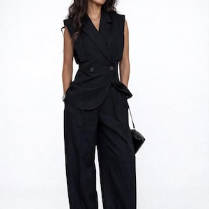 May include: A black sleeveless blazer and wide-leg trousers suit. The blazer has a double-breasted design with two buttons and a draped lapel. The trousers are high-waisted and loose-fitting. A small black handbag hangs from the shoulder.