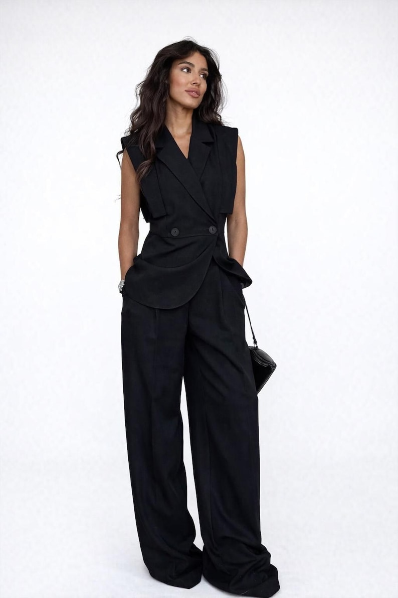May include: A black sleeveless blazer and wide-leg trousers suit. The blazer has a double-breasted design with two buttons and a draped lapel. The trousers are high-waisted and loose-fitting. A small black handbag hangs from the shoulder.