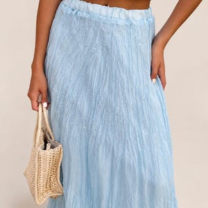 May include: A light blue maxi skirt with a crinkled, textured fabric finish and an elasticized waistband, paired with a small, beige woven straw handbag featuring two top handles and a structured, rectangular base design.