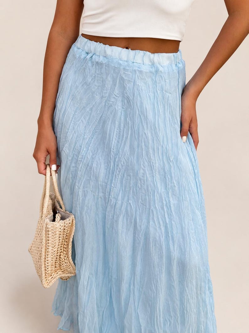 May include: A light blue maxi skirt with a crinkled, textured fabric finish and an elasticized waistband, paired with a small, beige woven straw handbag featuring two top handles and a structured, rectangular base design.