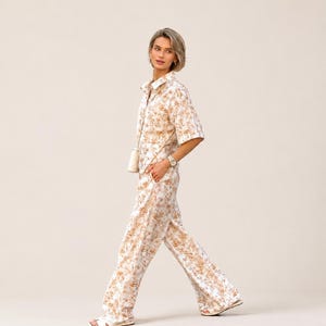 Puede incluir: Un conjunto de dos piezas a juego con una camisa con botones y pantalones de pernera ancha. El conjunto es blanco con un estampado floral repetido en tonos marrones y beige. La persona lleva un reloj y sandalias.