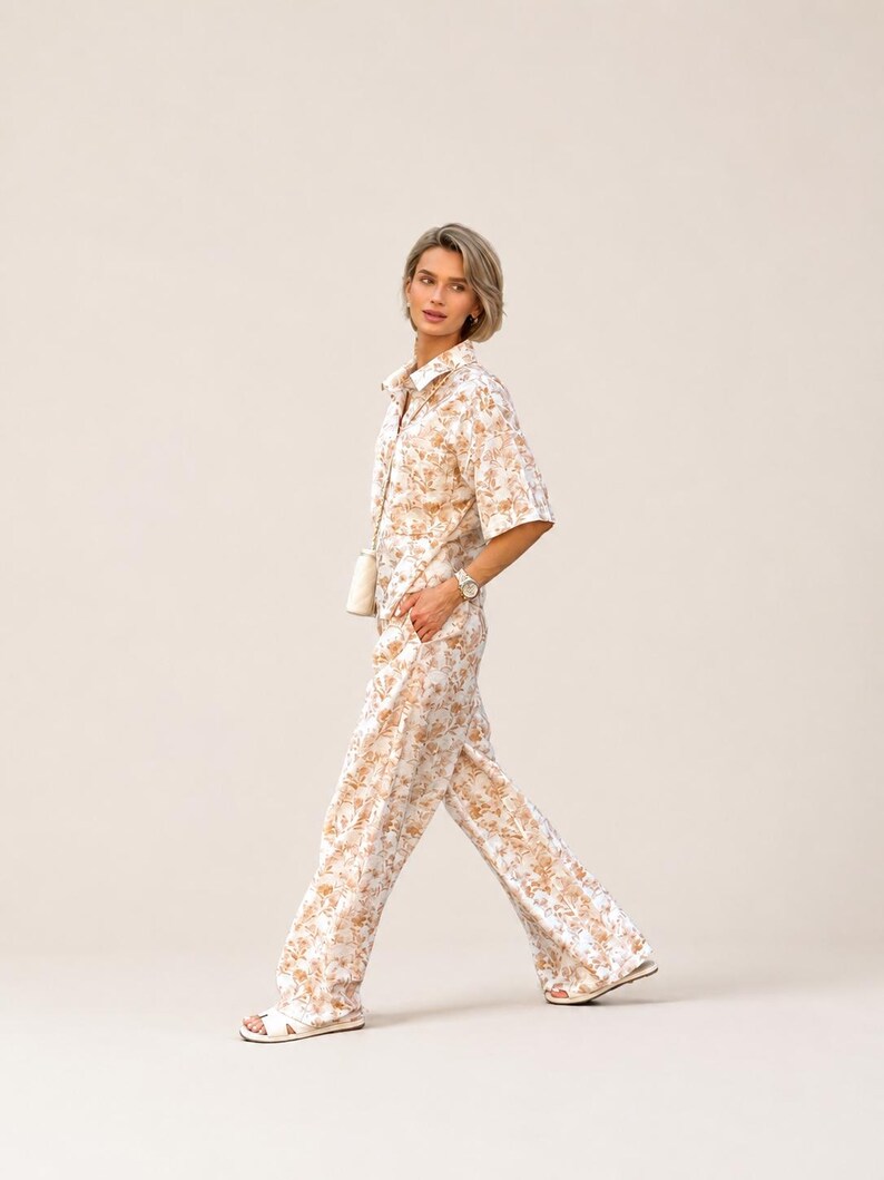 Puede incluir: Un conjunto de dos piezas a juego con una camisa con botones y pantalones de pernera ancha. El conjunto es blanco con un estampado floral repetido en tonos marrones y beige. La persona lleva un reloj y sandalias.