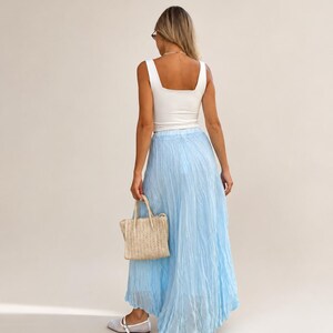 May include: A white sleeveless tank top paired with a long, light blue crinkled maxi skirt. The outfit is accessorized with a small, woven beige handbag featuring two top handles and light-colored flat shoes with a single strap across the top.