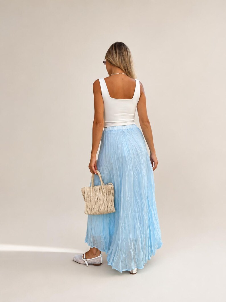 May include: A white sleeveless tank top paired with a long, light blue crinkled maxi skirt. The outfit is accessorized with a small, woven beige handbag featuring two top handles and light-colored flat shoes with a single strap across the top.