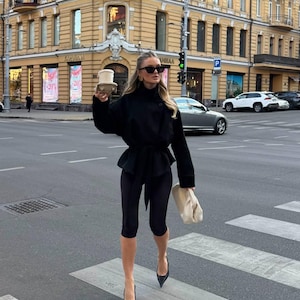 May include: A black long-sleeved jacket with a belted waist, black cropped leggings, black pointed-toe heels, and a cream-colored gathered clutch bag. The outfit is accessorized with dark-tinted sunglasses and a disposable coffee cup in a holder.
