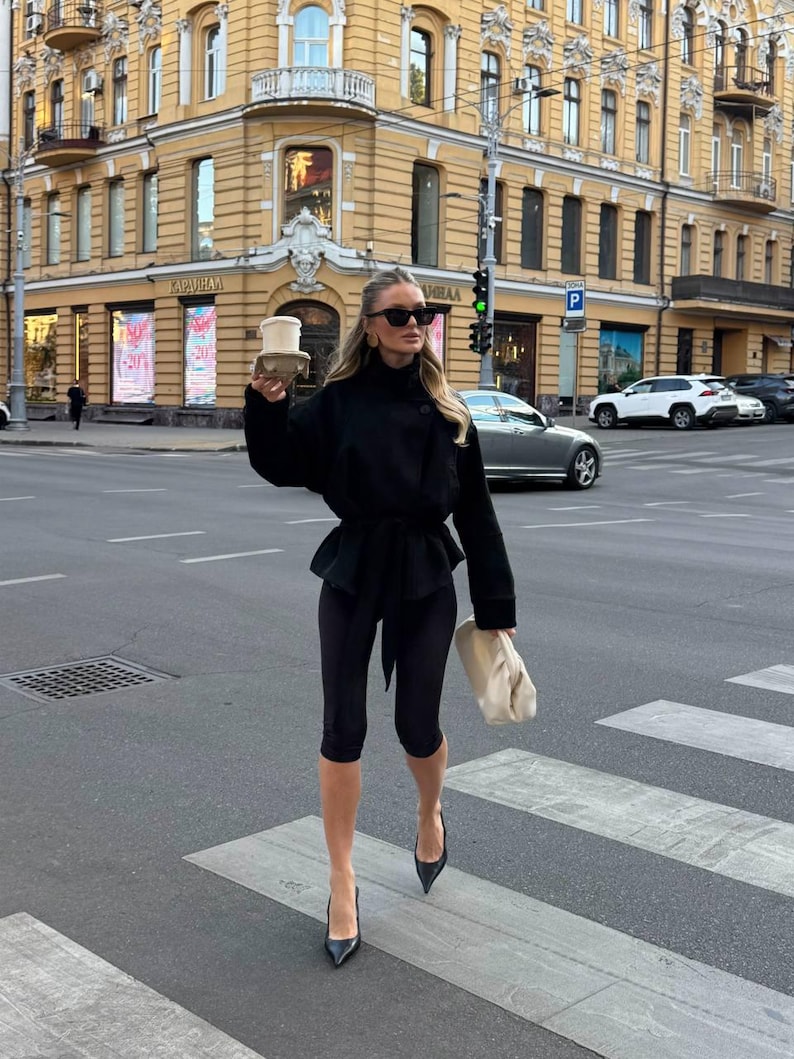May include: A black long-sleeved jacket with a belted waist, black cropped leggings, black pointed-toe heels, and a cream-colored gathered clutch bag. The outfit is accessorized with dark-tinted sunglasses and a disposable coffee cup in a holder.