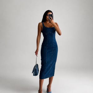 May include: A dark blue denim midi dress with a square neckline, thick shoulder straps, and vertical seam detailing along the front. The dress is paired with light blue denim open-toe heeled mules and a small matching dark blue denim handbag.