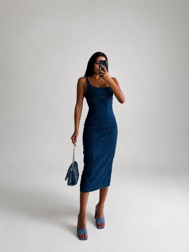 May include: A dark blue denim midi dress with a square neckline, thick shoulder straps, and vertical seam detailing along the front. The dress is paired with light blue denim open-toe heeled mules and a small matching dark blue denim handbag.