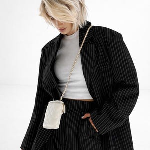 May include: A woman wearing a black pinstripe suit with a white ribbed tank top. A small, white quilted crossbody bag hangs from a gold chain. The woman is holding a smartphone.