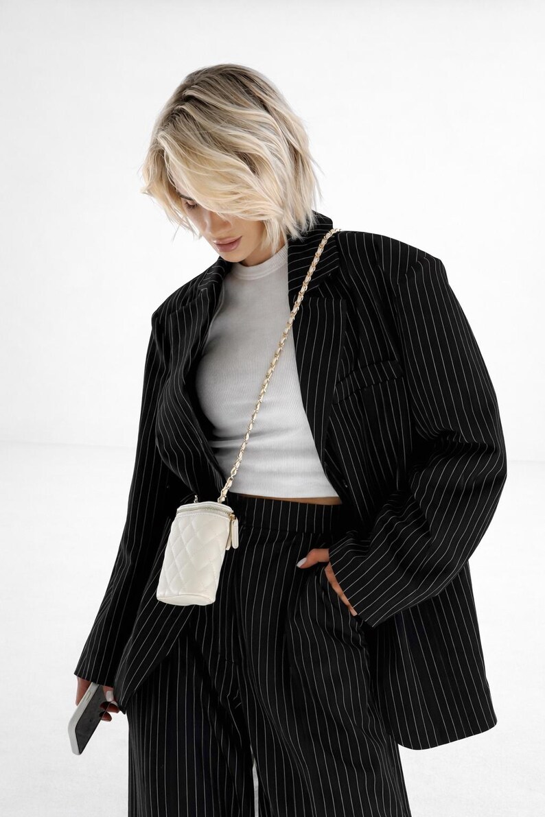 May include: A woman wearing a black pinstripe suit with a white ribbed tank top. A small, white quilted crossbody bag hangs from a gold chain. The woman is holding a smartphone.