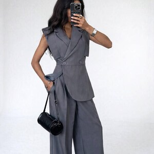 May include: A grey sleeveless blazer and wide-leg trousers suit. The blazer features a double-breasted design with an asymmetrical cut. A black cylindrical handbag hangs from the left shoulder. The outfit is made of a smooth, solid-coloured fabric.