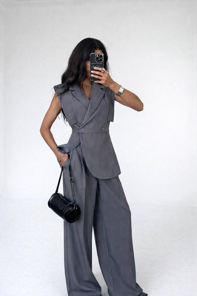 May include: A grey sleeveless blazer and wide-leg trousers suit. The blazer features a double-breasted design with an asymmetrical cut. A black cylindrical handbag hangs from the left shoulder. The outfit is made of a smooth, solid-coloured fabric.