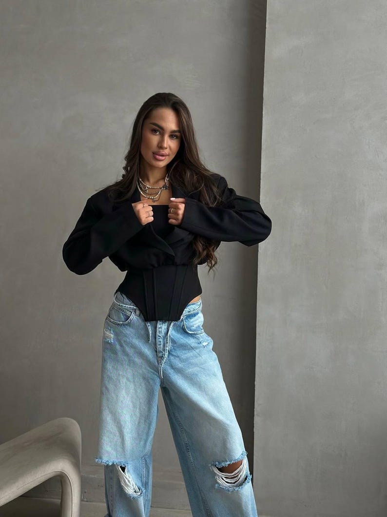 May include: A black blazer and corset top paired with light blue ripped jeans. The outfit is accessorized with layered necklaces. The model is standing in front of a gray wall.