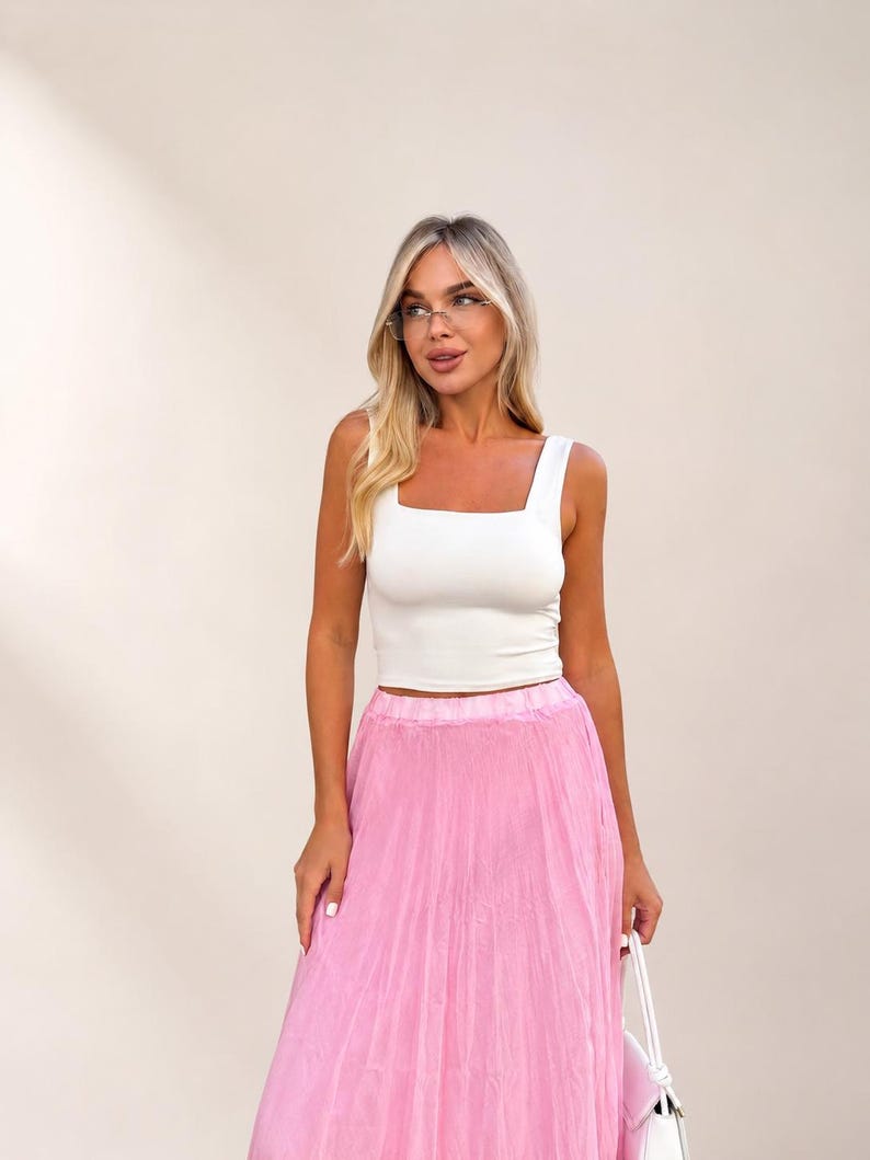 May include: A white sleeveless crop top with a square neckline paired with a long, flowing pink pleated skirt. The outfit is completed by a small white handbag held at the side, creating a coordinated ensemble with a clean and simple aesthetic.