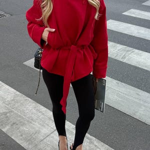 May include: A bright red wrap jacket with a high collar and a matching tie belt at the waist, paired with black stirrup leggings and black pointed-toe heels. A black quilted shoulder bag with a gold chain strap is visible on the side.