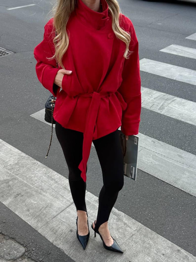 May include: A bright red wrap jacket with a high collar and a matching tie belt at the waist, paired with black stirrup leggings and black pointed-toe heels. A black quilted shoulder bag with a gold chain strap is visible on the side.