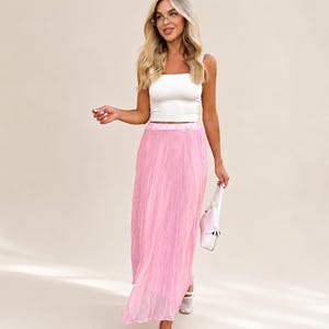 May include: A white sleeveless crop top with a square neckline paired with a long, pleated, light pink skirt. The outfit is accessorized with a small, light pink handbag and white mesh-style flat shoes, creating a coordinated ensemble.