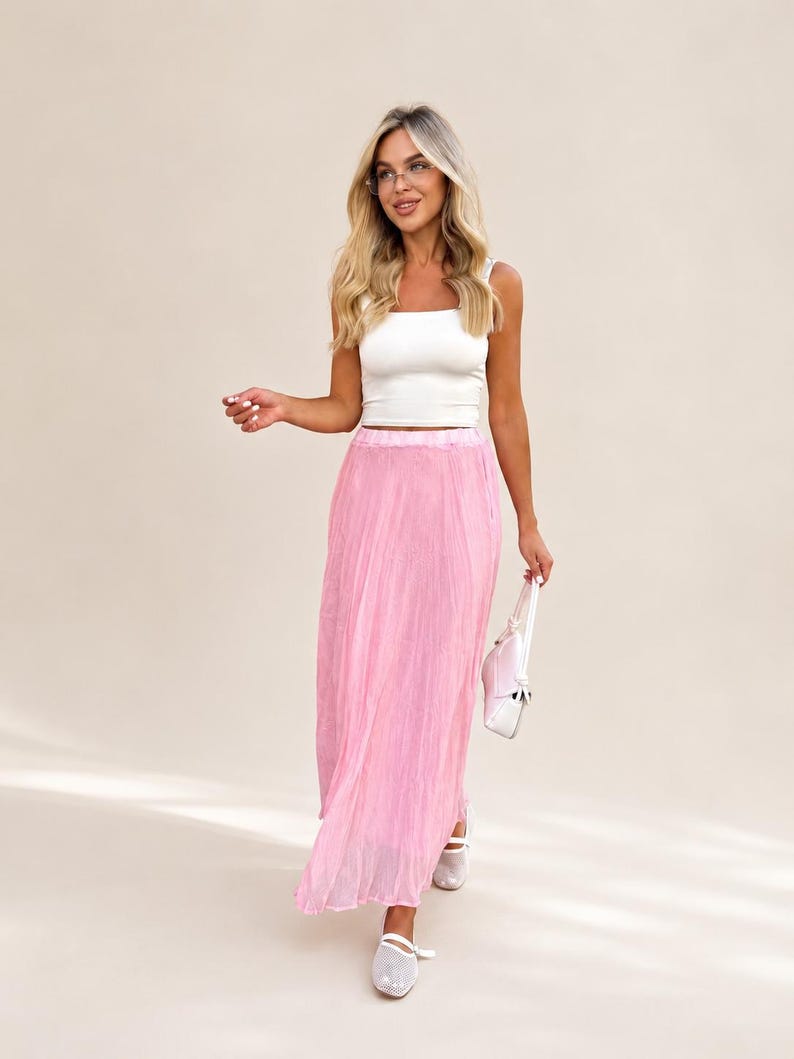 May include: A white sleeveless crop top with a square neckline paired with a long, pleated, light pink skirt. The outfit is accessorized with a small, light pink handbag and white mesh-style flat shoes, creating a coordinated ensemble.