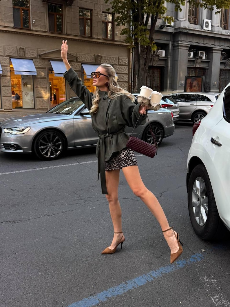 May include: An olive green belted jacket with a high collar, a leopard print mini skirt, brown pointed-toe heels with ankle straps, a dark burgundy cylindrical shoulder bag, and oversized brown sunglasses, while holding two white coffee cups.