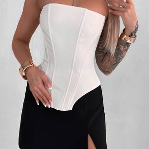 May include: A white strapless corset top with vertical seams is paired with a black mini skirt featuring a side slit. The outfit is accessorized with gold bracelets.