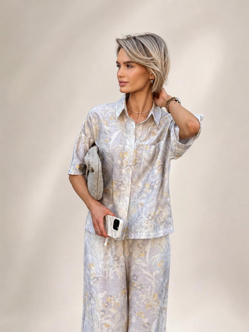 Puede incluir: Un conjunto de dos piezas a juego con una camisa con botones y pantalones de pernera ancha. El conjunto est&aacute; adornado con un estampado floral en tonos azul claro, blanco y amarillo. Un peque&ntilde;o clutch texturizado y una funda blanca para tel&eacute;fono completan el conjunto.