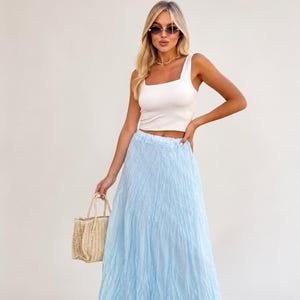 May include: A white sleeveless crop top with a square neckline paired with a long, light blue, crinkled texture maxi skirt. A small, beige, woven straw handbag with two handles is held at the side. The outfit is completed with dark sunglasses.