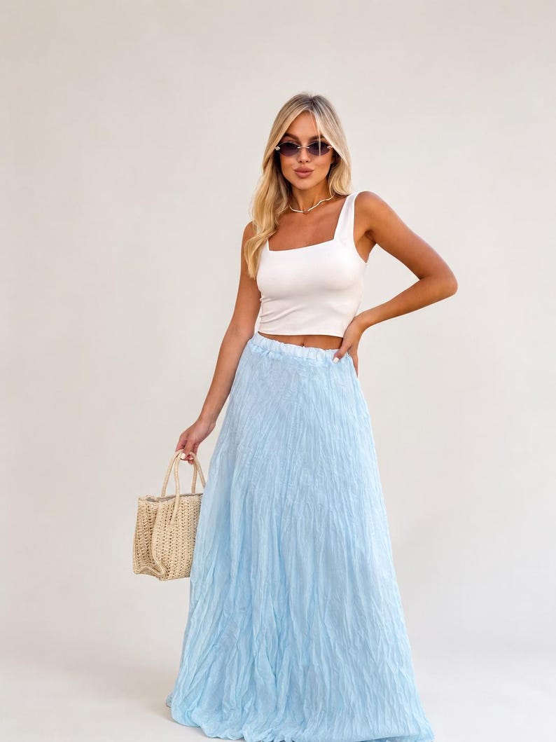 May include: A white sleeveless crop top with a square neckline paired with a long, light blue, crinkled texture maxi skirt. A small, beige, woven straw handbag with two handles is held at the side. The outfit is completed with dark sunglasses.