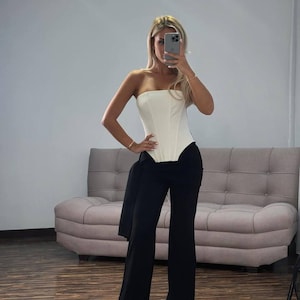 May include: A strapless white corset top paired with high-waisted, wide-leg black pants. A black jacket is draped over the pants. The outfit is worn in a room with a light-colored sofa and a wooden floor.