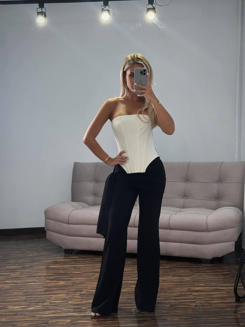May include: A strapless white corset top paired with high-waisted, wide-leg black pants. A black jacket is draped over the pants. The outfit is worn in a room with a light-colored sofa and a wooden floor.