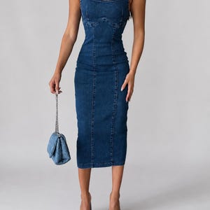 May include: A dark blue denim midi dress featuring a square neckline, thick shoulder straps, and vertical seam detailing along the bodice and skirt. The dress is paired with a matching quilted denim handbag with a silver chain strap and blue denim mules.