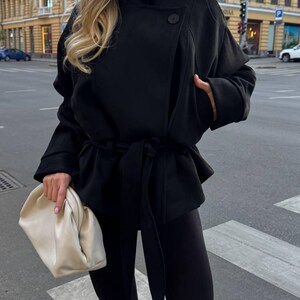 May include: A black jacket with a high collar, two visible buttons, and a tie-waist belt, paired with black leggings and a cream-colored, soft-structured clutch bag. The jacket features long sleeves and a wrap-style front closure.