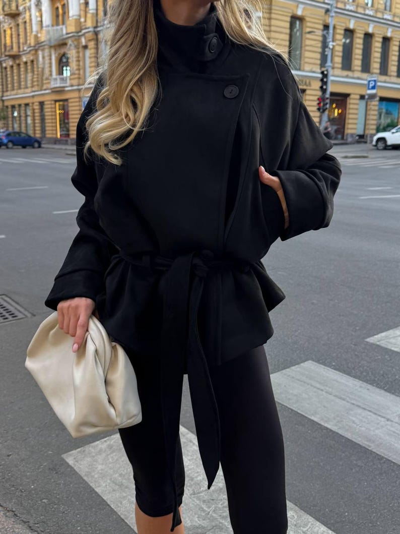 May include: A black jacket with a high collar, two visible buttons, and a tie-waist belt, paired with black leggings and a cream-colored, soft-structured clutch bag. The jacket features long sleeves and a wrap-style front closure.