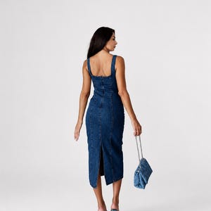 May include: A dark blue denim midi dress featuring a square neckline, thick shoulder straps, and a vertical seam construction. The dress has a back slit at the hem and is paired with a matching quilted denim handbag with a silver chain strap.