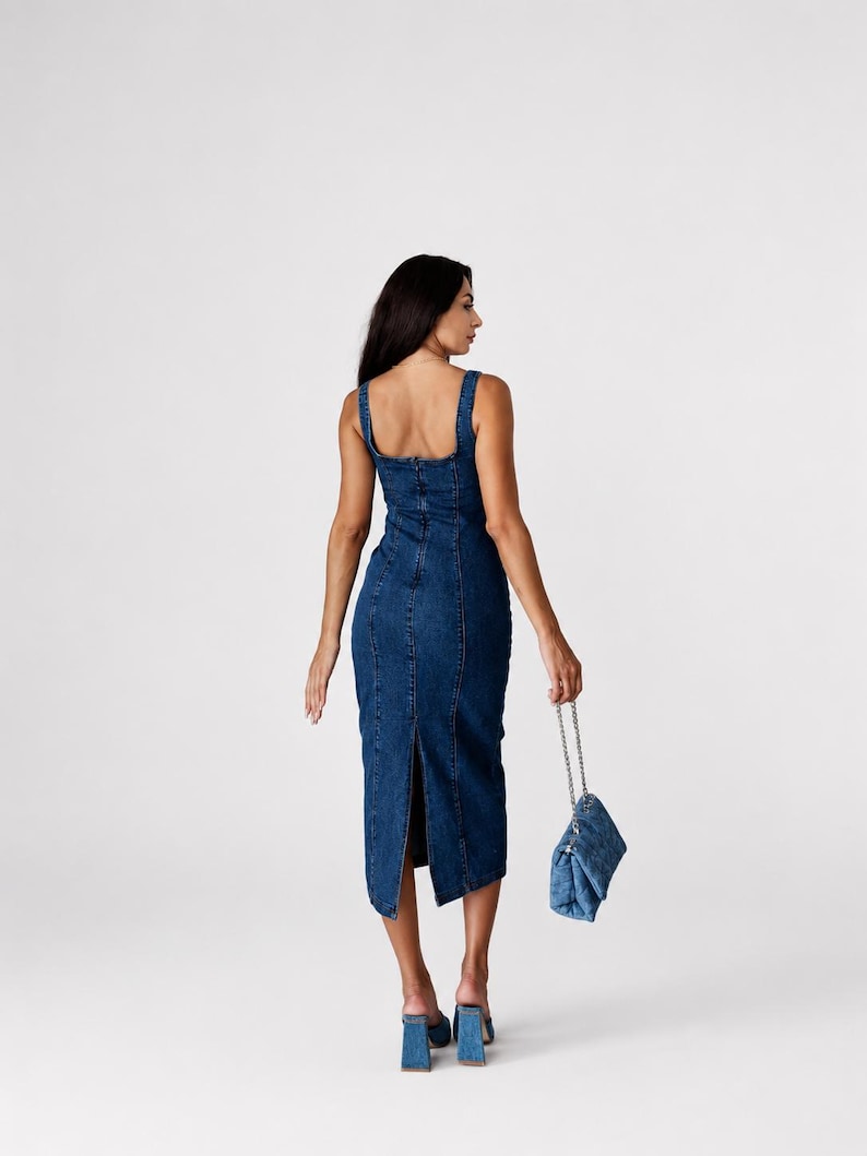 May include: A dark blue denim midi dress featuring a square neckline, thick shoulder straps, and a vertical seam construction. The dress has a back slit at the hem and is paired with a matching quilted denim handbag with a silver chain strap.