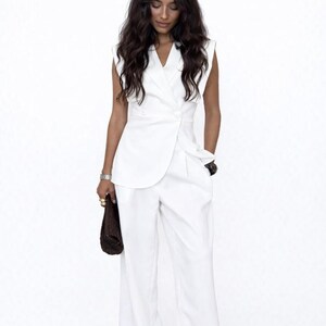 May include: A white sleeveless blazer and wide-leg trousers. The blazer features a double-breasted design with two buttons. The outfit is a monochromatic ensemble, suitable for formal or business wear.
